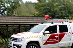 Tetőfóliázás a háttérben, a kép előterében egy mentőszervezeti autó.